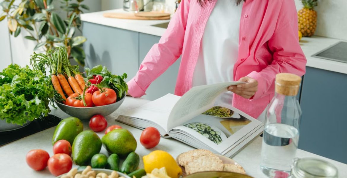 A person browsing a cookbook surrounded by fresh vegetables in a bright kitchen setting.