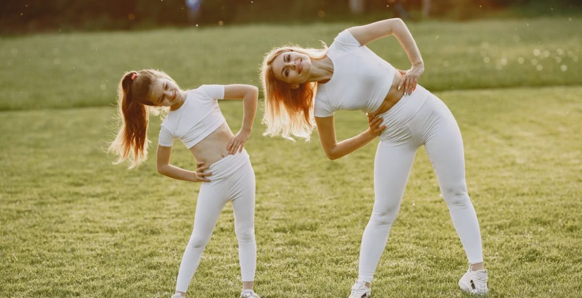 A mother and daughter in activewear stretching on a sunny day in a grassy field.