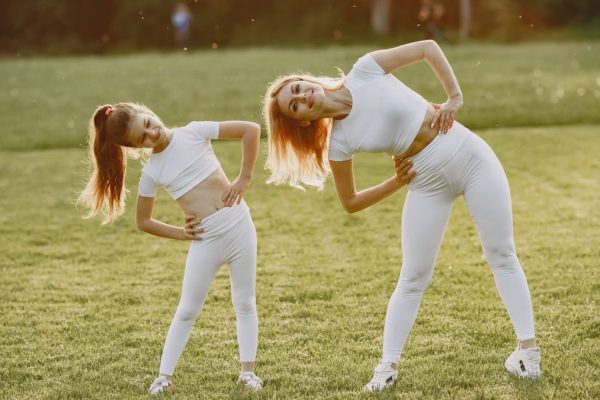 A mother and daughter in activewear stretching on a sunny day in a grassy field.