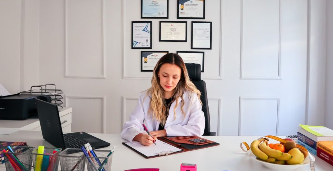 Nutritionist sits at a modern office desk writing, with fruits and diplomas displayed, emphasizing health and professionalism.